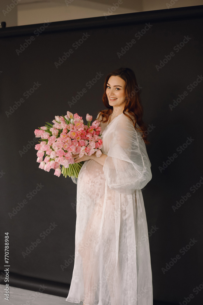 Portrait of attractive pregnant woman in white transparent dress with bouquet of pink tulips on black background. Stylish pregnant woman. Portrait of attractive pregnant woman in white transparent dress with bouquet of pink tulips on black background. Stylish pregnant woman.