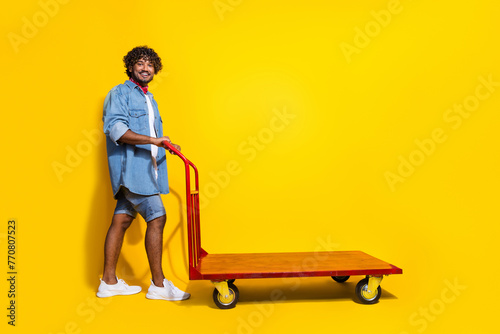 Full length photo of handsome young guy push cart warehouse wear trendy denim garment red scarf isolated on yellow color background