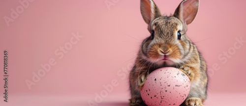 The image beautifully captures a brown rabbit with large eyes holding onto a single speckled pink Easter egg against a pink background