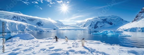 a group of penguins frolicking on the snow-covered ground against the backdrop of majestic blue mountains, tranquil waters dotted with floating icebergs, and the radiant sun shining brightly