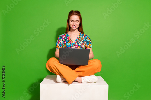 Full size photo of pretty young woman sit cube use laptop wear top isolated on bright green color background photo