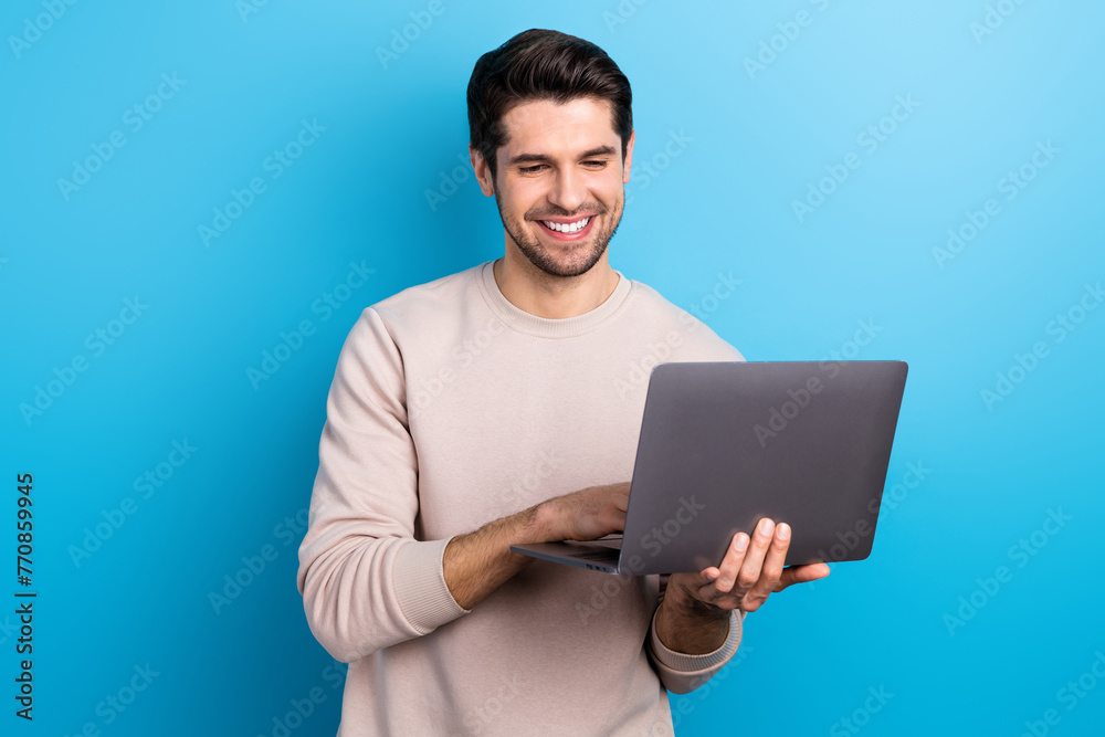 Portrait of satisfied pleasant guy with brunet hair wear light pullover look at laptop typing message isolated on blue color background