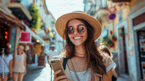 A smiling tourist in a sun hat and sun glasses is holding a phone in her hand. Summer day, background of an old European city