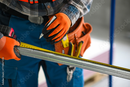 a construction worker in overalls, gloves and a tool belt measures the length of a plasterboard profile with a meter - close-up view photo