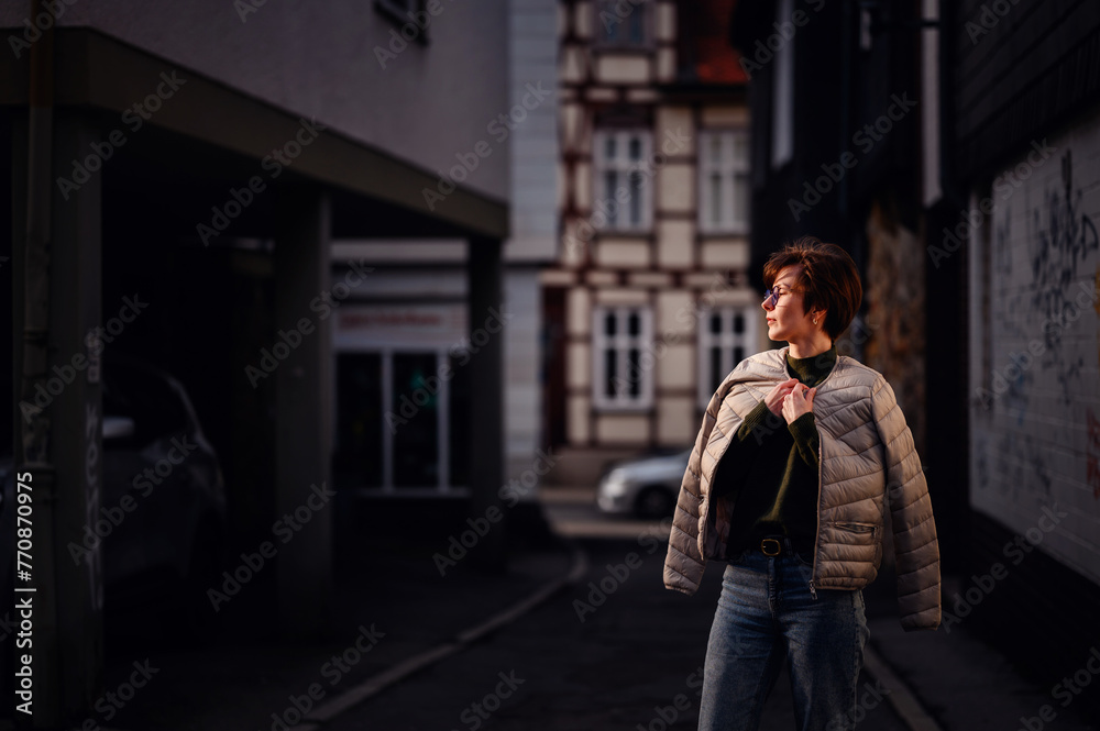 Fototapeta premium A fashion-forward woman with glasses and a green sweater carries her jacket confidently in a dimly lit urban alley.