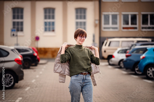 A spirited young woman with a radiant smile is caught in the act of slipping on a light jacket over her green sweater, outdoors in a parking area. © Иванна Емельянова