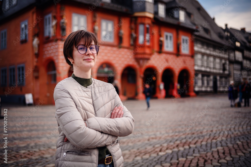 A woman stands confidently in a historic town square, her arms crossed, with traditional architecture creating a charming backdrop.