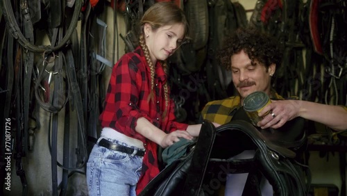 Father and daughter owners of a horse ranch, clean and apply ointment to protect leather to a saddle in the workshop