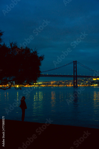 Lisbon April 25 bridge on a cloudy day blue hour, a person and tree silhouette lights on