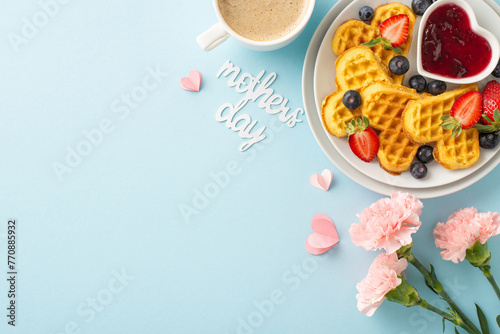 Mother's Day breakfast display: top view heart-shaped waffles, raspberries, blueberries, cup of drink, fresh flowers, card "mothers day," on a pastel blue background, space for text