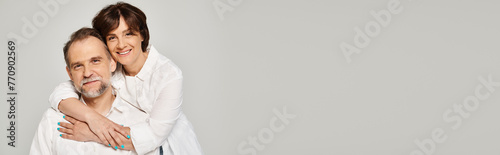 Portrait of middle aged smiling couple hugging and looking at camera on grey background, banner photo