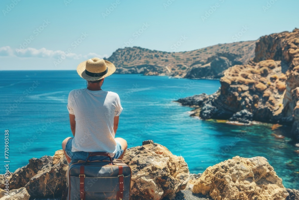 A traveler with a straw hat sitting on a suitcase looks towards a serene blue sea against a rocky coastline