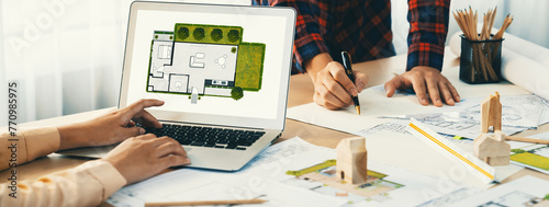 Professional architect represented ECO house displayed on laptop to project manager on messy table with wooden block, blueprint, architectural document placed at modern office. Close up. Delineation. photo