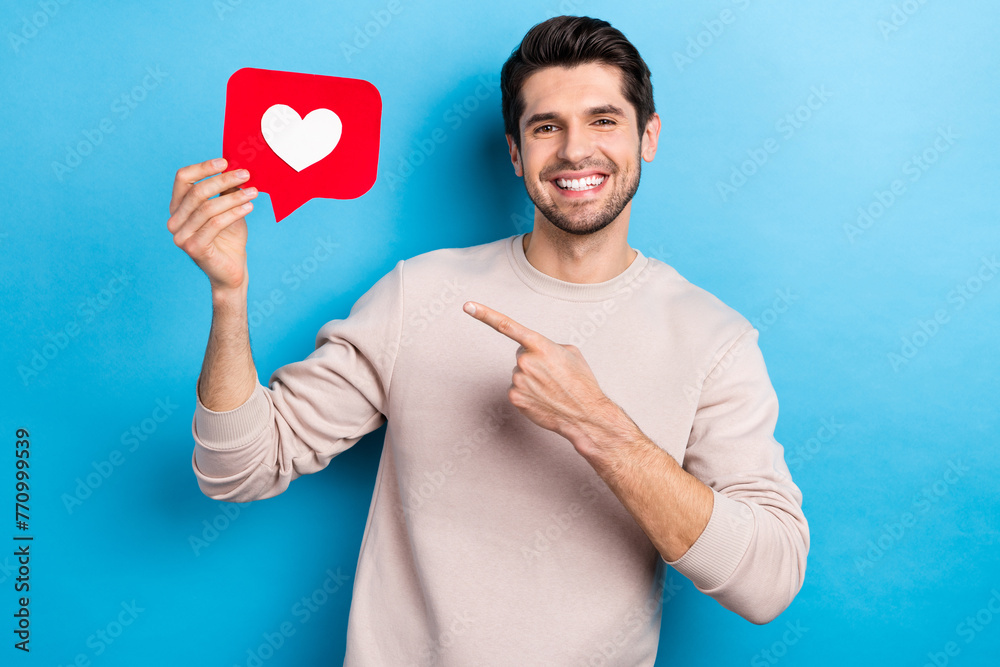 Photo of positive toothy beaming man stylish haircut dressed beige sweatshirt directing at like icon isolated on blue color background Photo of positive toothy beaming man stylish haircut dressed beige sweatshirt directing at like icon isolated on blue color background