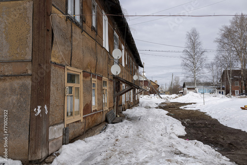 An old house in a provincial town in Russia. Peeling facade, old windows, satellite TV, thawed snow. Greay sky. In the distance, a banner of an electrical equipment store. © PuzzleSoul