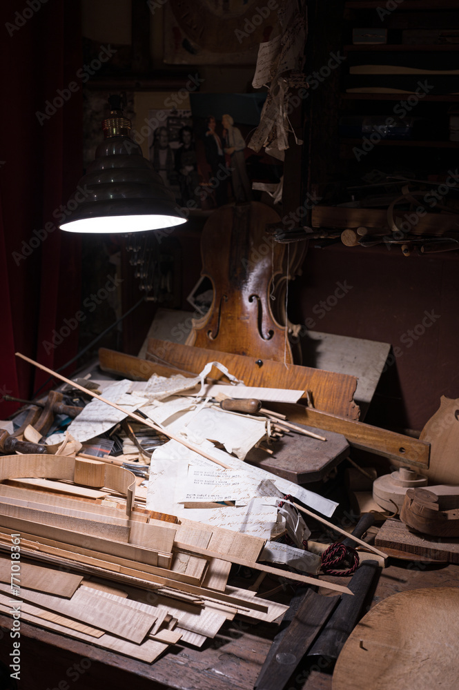 Detail of the messy work table of the master craftsman luthier maker of high-end violins. pieces of wood, papers, notes, tools in a dark workshop illuminated by a lamp