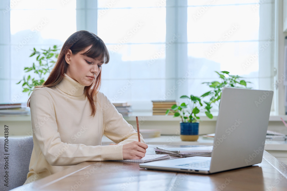 College student girl sitting at desk using computer, making notes in notebook College student girl sitting at desk using computer, making notes in notebook