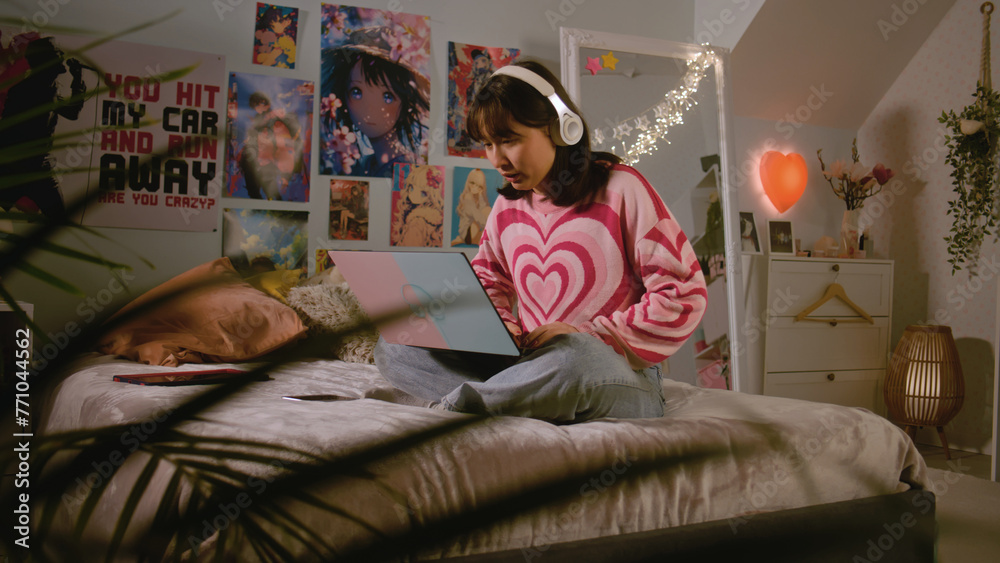 Obraz premium Asian teenage girl in headphones listens to music, chats and surfs the Internet on laptop while sitting on the bed in her bedroom. Girl spending leisure time and having fun at home. Lifestyle concept.