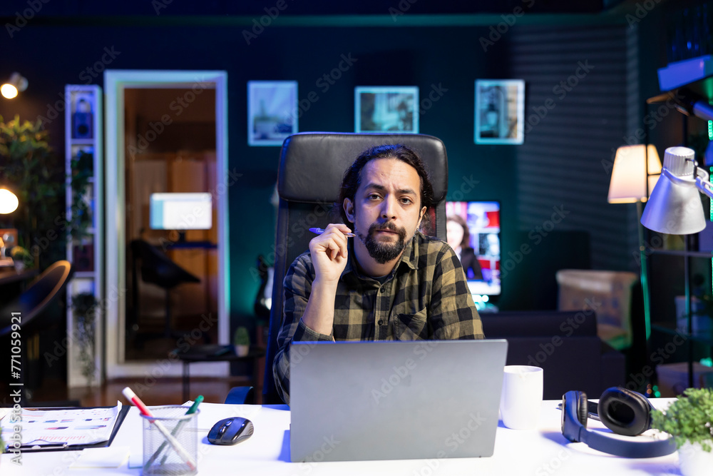 Portrait of young man sitting in front of his laptop at his home desk, working from home. Serious male individual holding a pen while looking at the camera, preparing for work and research. Portrait of young man sitting in front of his laptop at his home desk, working from home. Serious male individual holding a pen while looking at the camera, preparing for work and research.