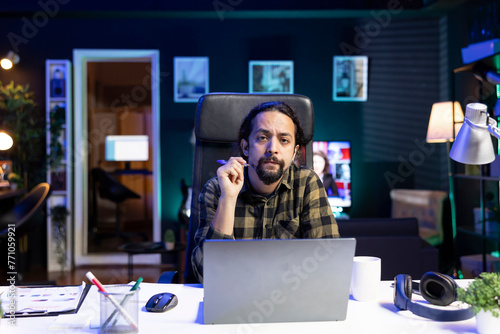 Portrait of young man sitting in front of his laptop at his home desk, working from home. Serious male individual holding a pen while looking at the camera, preparing for work and research.