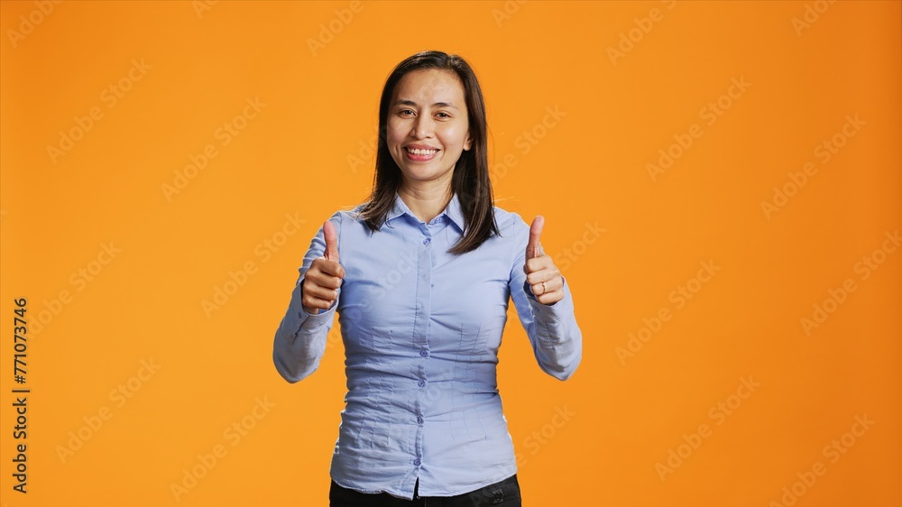 Asian model presenting thumbs up symbol in studio, expressing good vibes and success against orange background. Young smiling person giving like sign on camera, shows approval gesture. Asian model presenting thumbs up symbol in studio, expressing good vibes and success against orange background. Young smiling person giving like sign on camera, shows approval gesture.