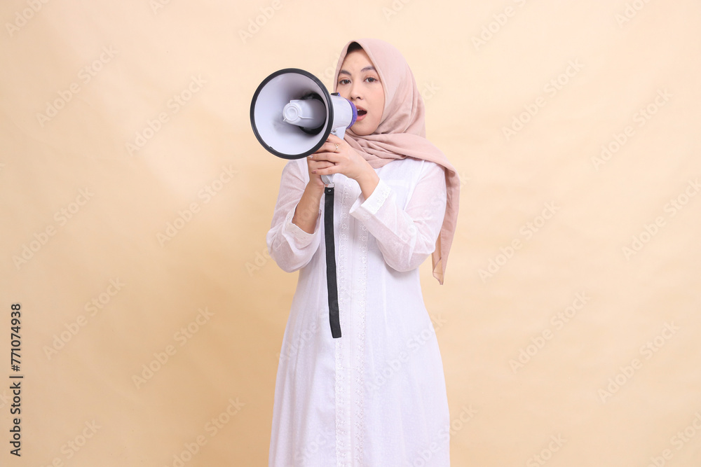 woman asian muslim wearing a hijab screams at the camera in surprise, holding a loudspeaker megaphone with both hands. Technology, broadcast and promotion concept woman asian muslim wearing a hijab screams at the camera in surprise, holding a loudspeaker megaphone with both hands. Technology, broadcast and promotion concept