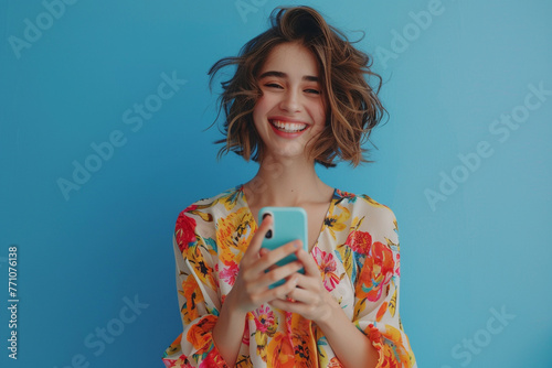 An alone, joyful, and exuberant woman is gesturing at her phone against a blue background. A cheerful female model is seen holding a cellphone and presenting a fresh, modern application concept.