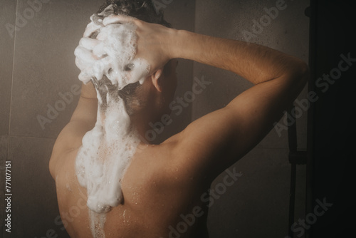 Back view of a young man lathering shampoo in his hair while taking a shower, depicting daily cleanliness routine.