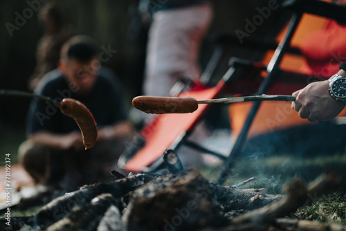 Close-up of sausages cooking on sticks over campfire with friends in the background enjoying outdoor camping fun