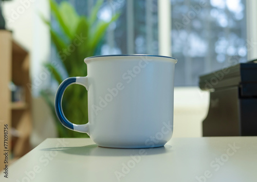A enamel white mug with a blue handle, a type of drinkware, is placed on a white table, a piece of tableware or serveware