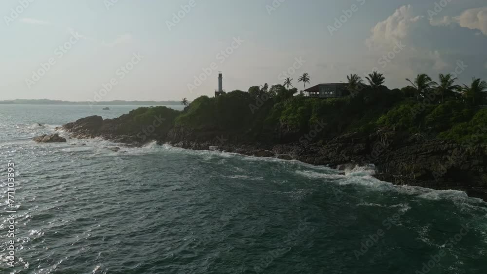 Drone view over ocean shows waves hitting rocky coast with hilltop lighthouse. Maritime beacon stands by sea for ship guidance, safety. Scenic seascape offers travel, adventure backdrop. Drone view over ocean shows waves hitting rocky coast with hilltop lighthouse. Maritime beacon stands by sea for ship guidance, safety. Scenic seascape offers travel, adventure backdrop.