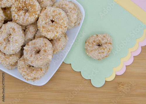 Cropped angle view of a square plate with round crumb donuts on a light wood table, one donut isolated on green paper napkin with pink and yellow napkins below. Scalloped edges on napkins.