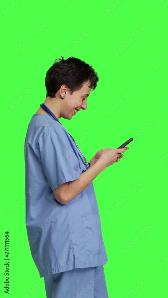 Profile Young nurse texting on smartphone against greenscreen backdrop, using mobile phone to check social media messages on work break. Medical assistant browsing online web pages. Camera B. Profile Young nurse texting on smartphone against greenscreen backdrop, using mobile phone to check social media messages on work break. Medical assistant browsing online web pages. Camera B.