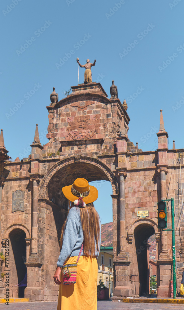 Young woman traveler in brown hat walks through of the Santa Clara Arch in San Francisco square. Cusco, Peru.