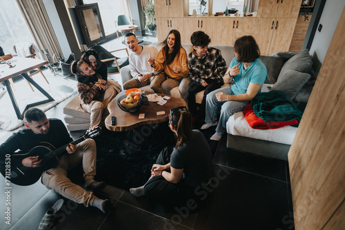 Schilderij op canvas Casual indoor gathering of friends sitting comfortably, playing guitar, and havi