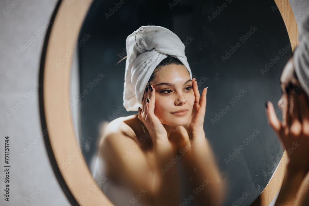 © qunica.com - A serene young woman performs a skincare routine, gently touching her face in front of a mirror in a cozy, dimly lit room.