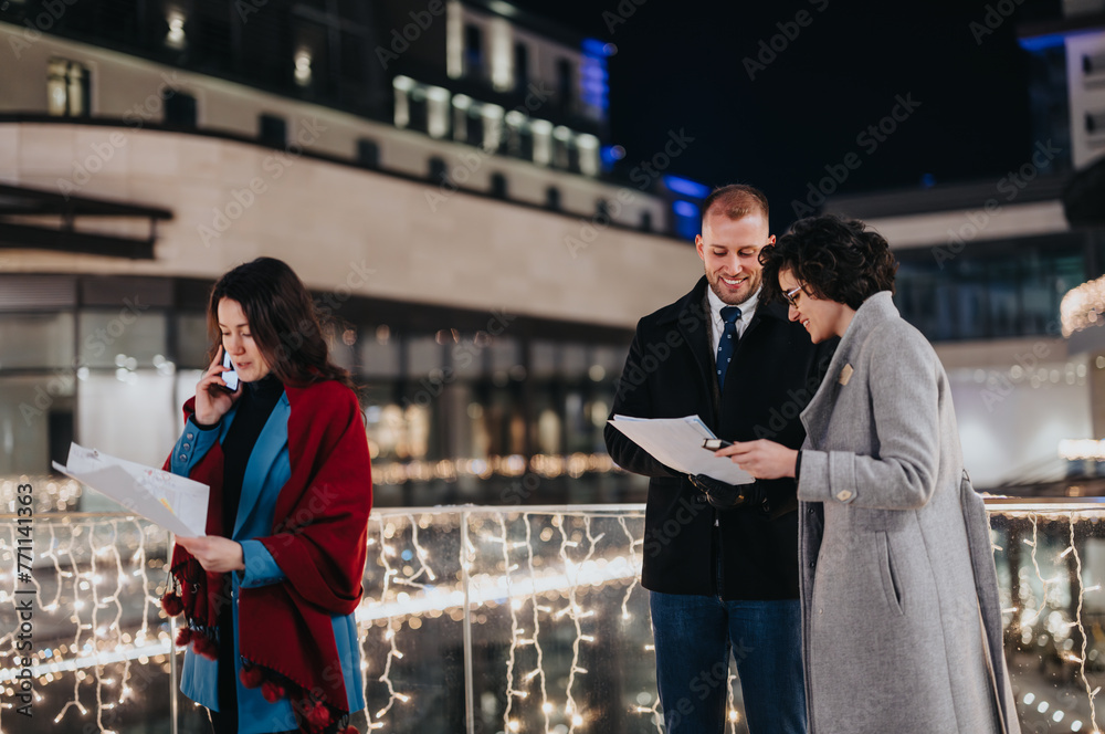 Naklejka premium Three professionals gathered outdoors at night during winter, discussing papers with festive lights in the background.