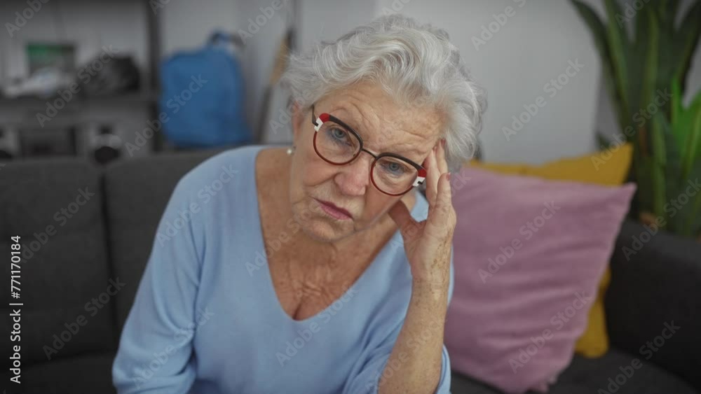 A pensive elderly woman with grey hair and glasses sits at home, concern evident in her expression. A pensive elderly woman with grey hair and glasses sits at home, concern evident in her expression.