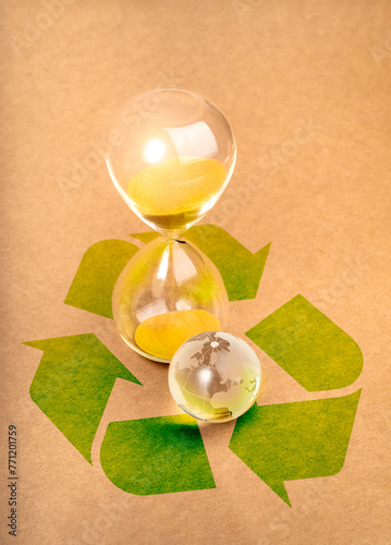 Environmental sustainability, time and responsibility, conserve resources concept. Small glass world globe near hourglass on big green recycle symbol on brown paper background, vertical style.