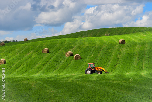 Rural country landscape with alfalfa field and cereal hay bales and tractor with fork loader in summer season
