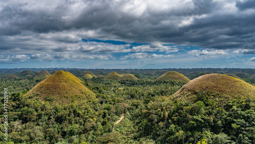 A miracle of nature - many amazing rounded Chocolate hills covered with brownish grass stretches to the horizon. A dirt road runs through a valley among lush tropical vegetation.Clouds in the blue sky