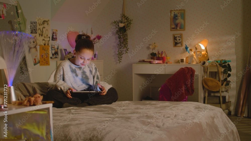 Young girl sits on bed in her room and surfs the internet or chats online using tablet. Cute schoolgirl spending leisure time at home scrolling social networks. Home with cozy and stylish interior.