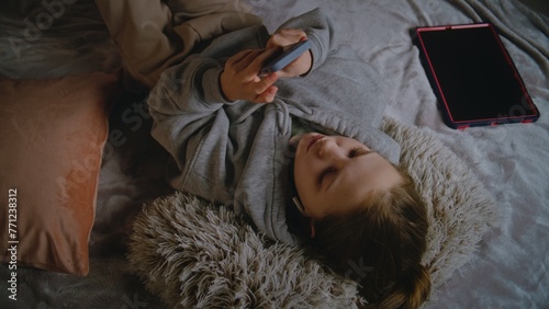 Young girl in headphones lies on the bed at home, listens to music and surfs the Internet using mobile phone. Caucasian teenager spends leisure time in comfort bedroom in daytime. Lifestyle concept.