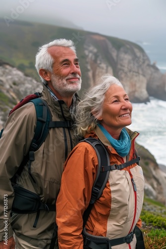Elderly senior smiling active couple with backpacks enjoying hiking and walking along coastline near mountains and sea or ocean, active retirement lifestyle filled with outdoor adventures.