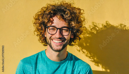 Candid portrait of a handsome curly-haired young man in a turquoise t-shirt and glasses on a yellow background laughing.