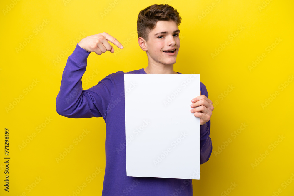 Teenager Russian man isolated on yellow background holding an empty placard with happy expression and pointing it Teenager Russian man isolated on yellow background holding an empty placard with happy expression and pointing it