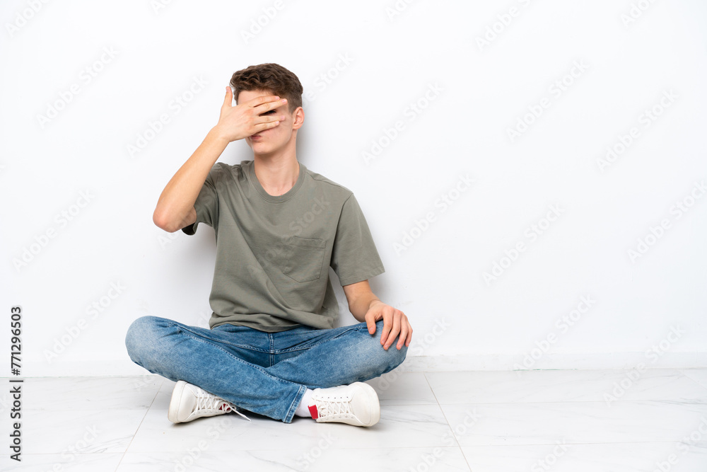 Teenager Russian man sitting on the floor isolated on white background covering eyes by hands and smiling Teenager Russian man sitting on the floor isolated on white background covering eyes by hands and smiling