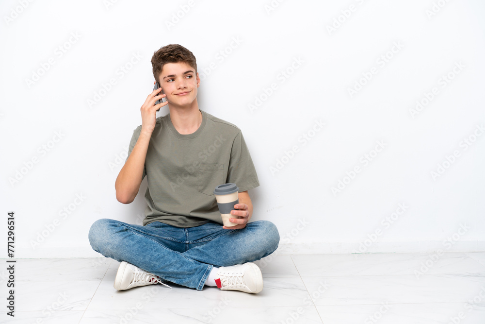 Teenager Russian man sitting on the floor isolated on white background holding coffee to take away and a mobile
