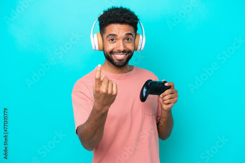 Young brazilian man playing with a video game controller isolated on blue background doing coming gesture