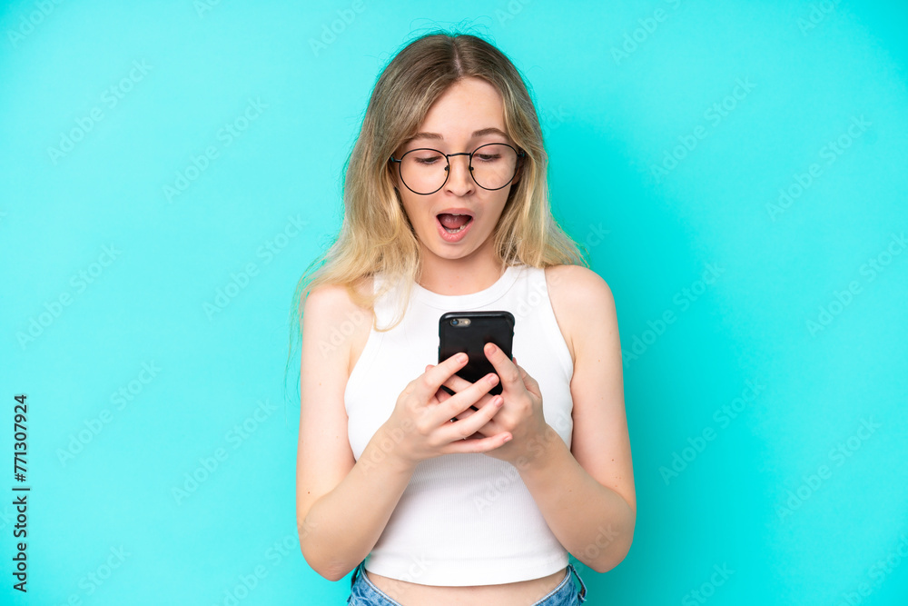 Blonde English young girl isolated on blue background looking at the camera while using the mobile with surprised expression Blonde English young girl isolated on blue background looking at the camera while using the mobile with surprised expression